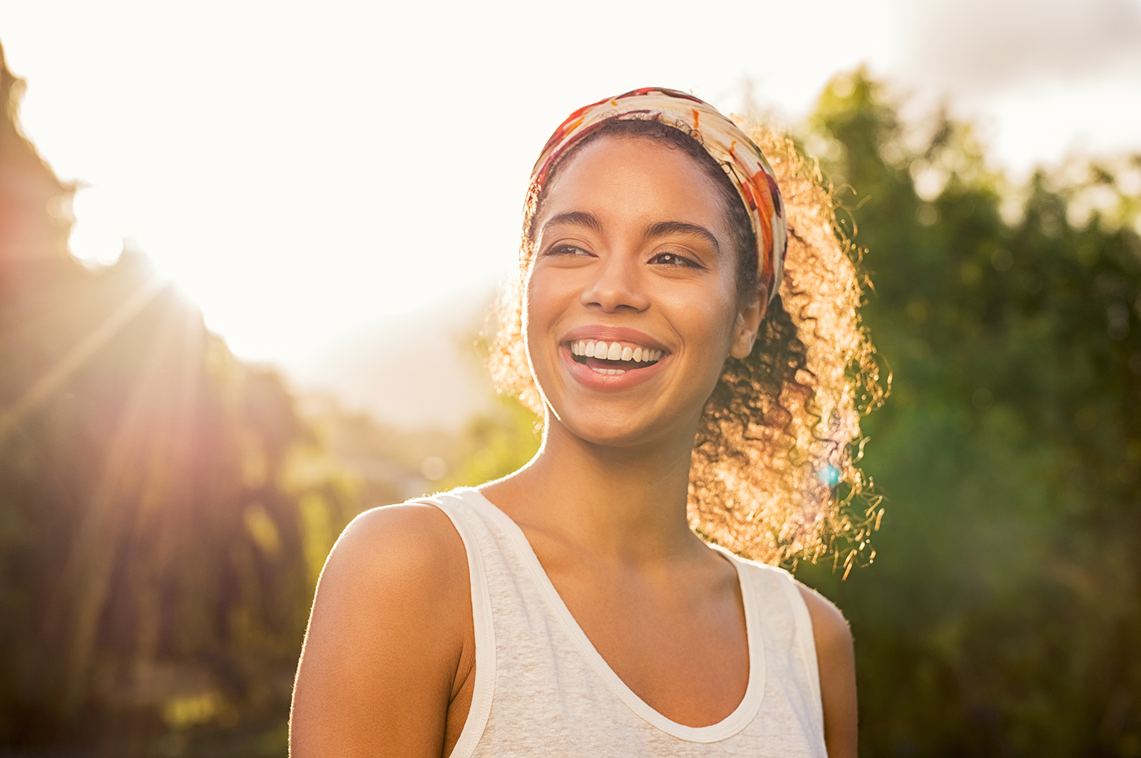 The image shows a smiling woman with curly hair wearing a patterned headscarf, standing outdoors during sunset with a bright sky background.