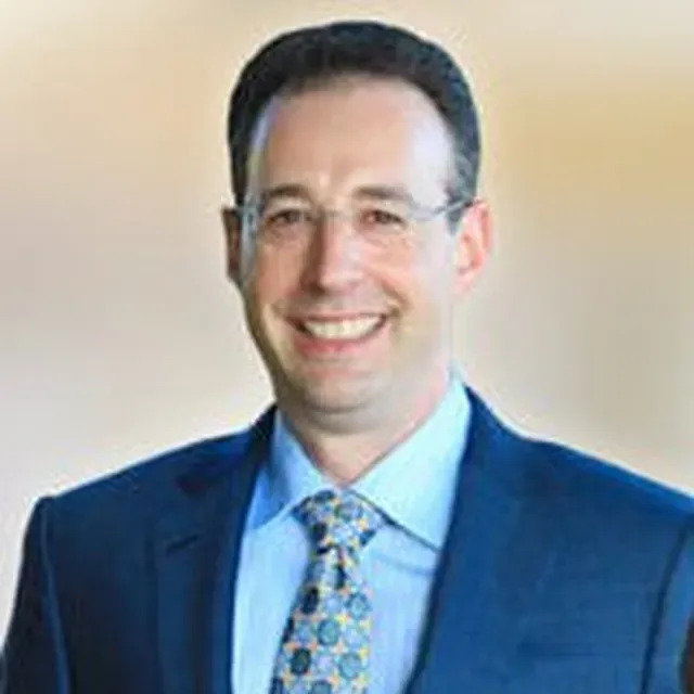 The image shows a man wearing glasses, smiling, and dressed in formal attire standing against a blurred background.