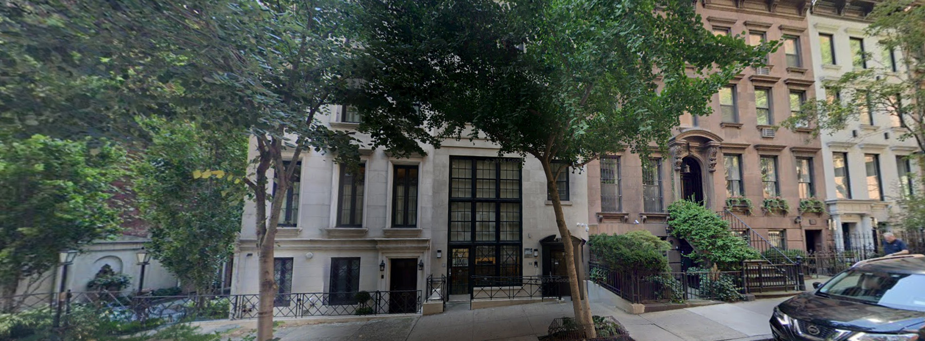 The image shows a residential street scene with a row of multi-story houses on one side, trees lining the sidewalks, and parked cars along the roadside.