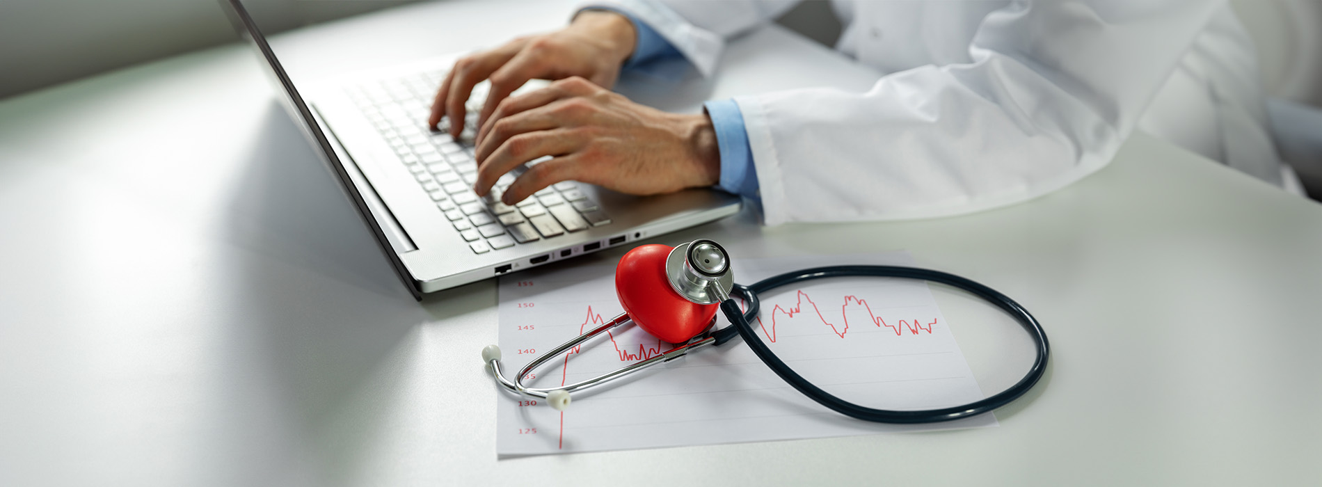 A person wearing a white coat with a stethoscope around their neck, using a laptop at a desk, with medical equipment and a red heart monitor next to them.