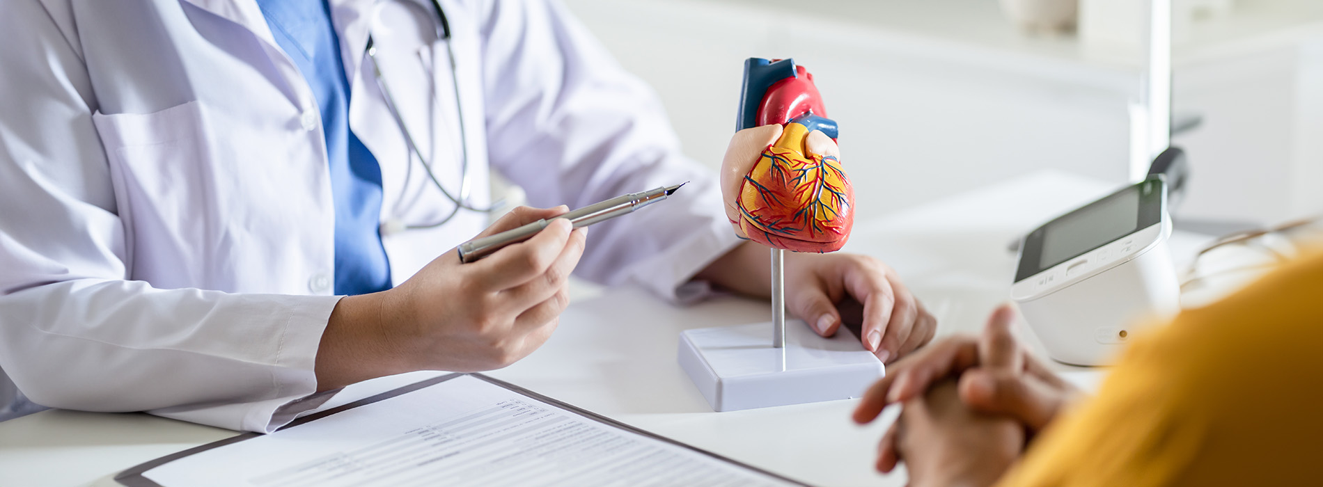 The image shows a medical professional sitting at a desk with a patient s file, wearing a stethoscope around their neck, and holding a tablet computer, while another person appears to be seated across from them in a consultation setting.