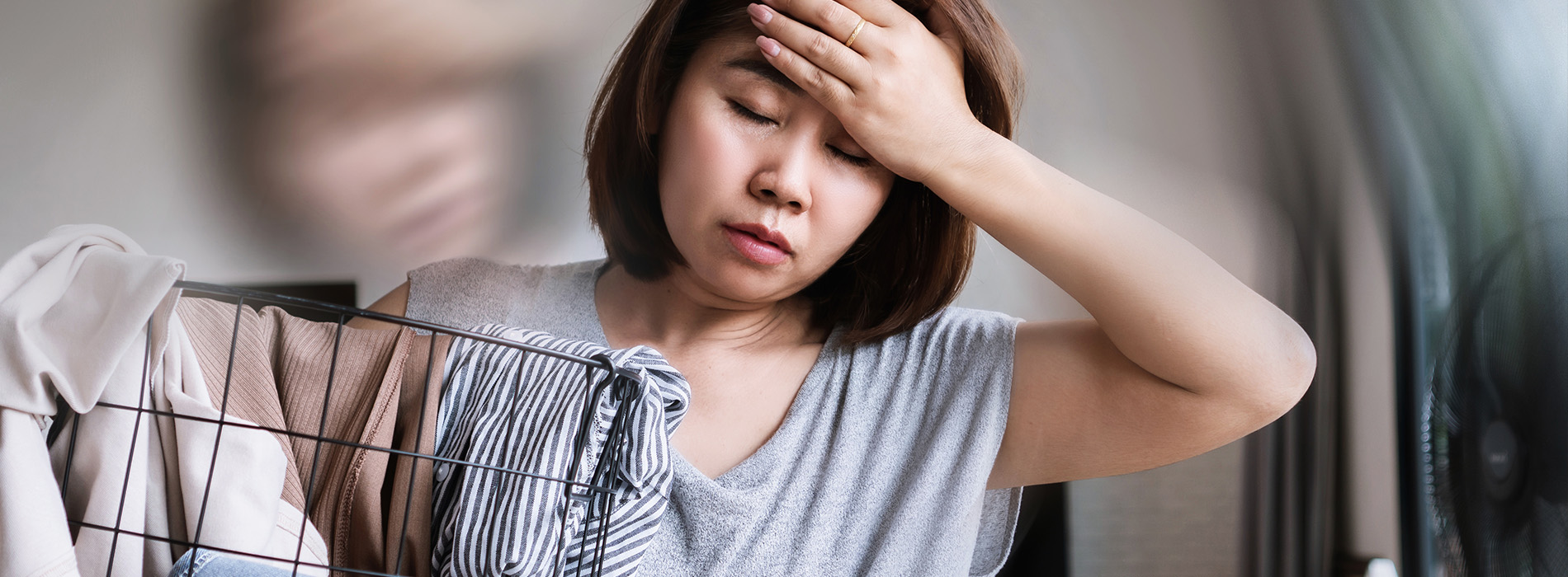 A woman with her hand on her forehead, possibly indicating discomfort or exhaustion, set against a blurred background with an ironing board visible.