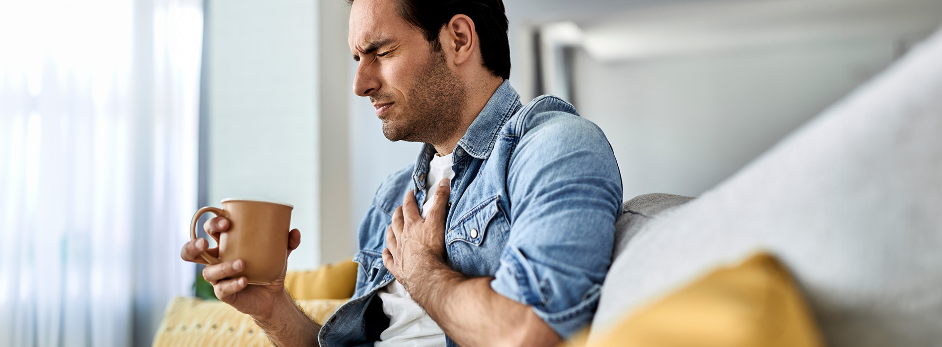 A man sitting on a couch with his hand on his chest, looking downwards while holding a cup.