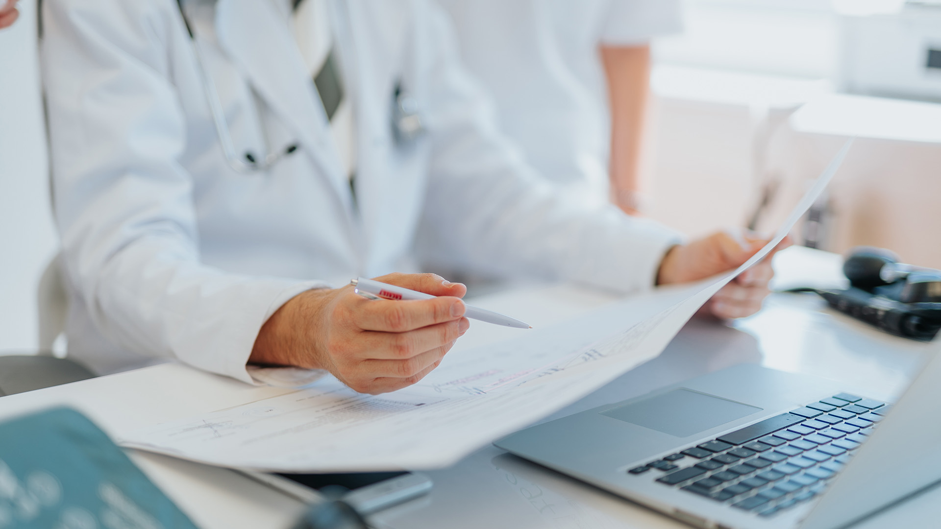 The image shows a medical professional sitting at a desk with a laptop, reviewing documents while wearing a white coat.