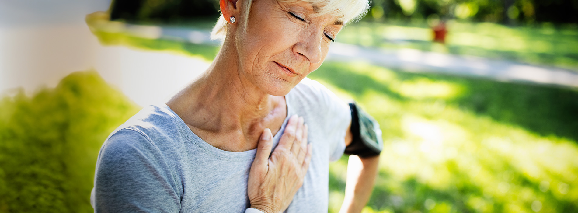The image shows an older woman with short hair wearing a gray top, standing outdoors on a sunny day, looking upwards, possibly towards the sky, with her hands clasped together near her chest.